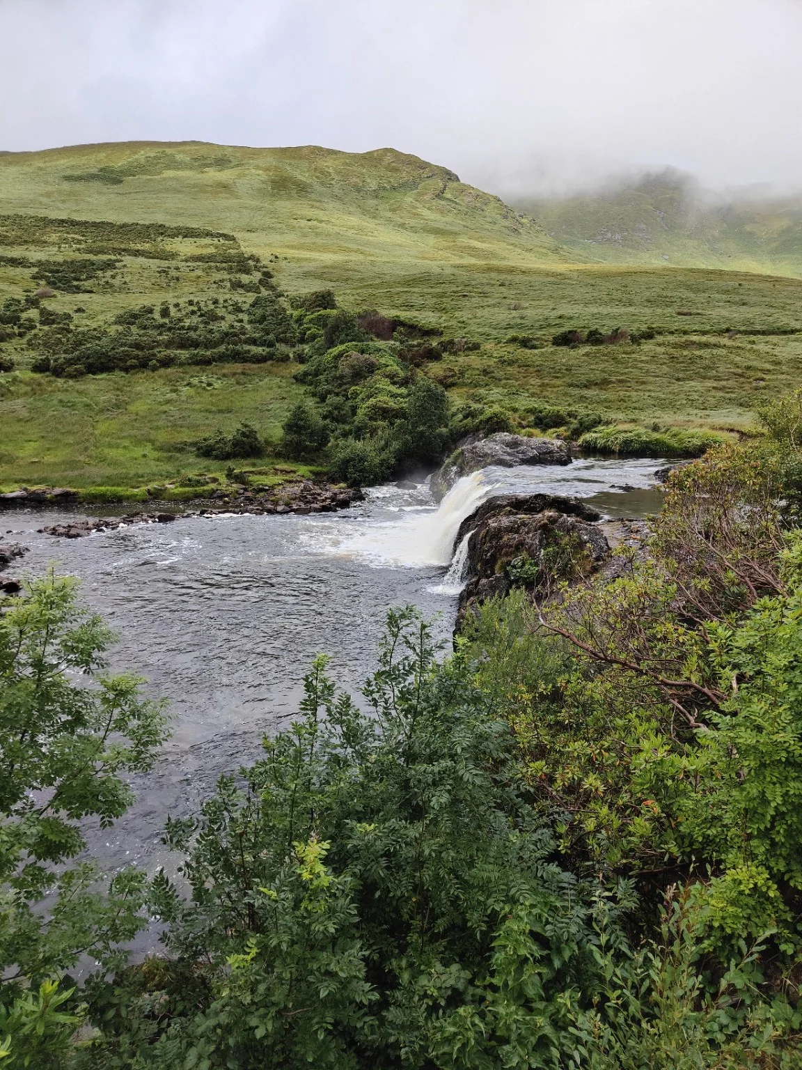 Een rivier stroomt over een rotsrand als een kleine waterval, omringd door groene hellingen en laaghangende wolken